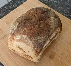 "Photograph of a large, pale-coloured lozenge-shaped bread loaf shown on a wooden tray"