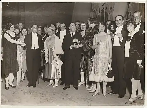 Large group photo of people standing, including white man in white tie and tails with performers and others either in day clothes or evening dress, some holding a drink and/or shaking hands