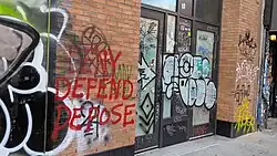 Graffiti reading "Deny Defend Depose" spraypainted with red paint on a brick wall in New York City.
