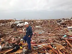 A man in blue clothes and a white hard hat walks up on a pile of debris from an EF5 tornado in 2013