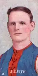 A brown-haired footballer in a sleeveless blue guernsey with vertical red stripes