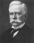 A headshot of a Caucasian male with a white handlebar moustache and white hair. He is wearing a white winged-collar shirt, black cravat and black coat.