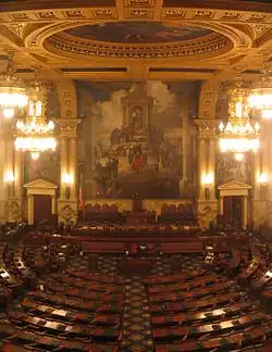 A view of a large, ornately-decorated room with several rows of curved desks arranged in a semicircle. A large mural is visible on the wall at the far end of the room.