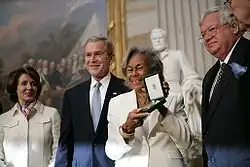 Black woman holding aloft award presented by President George W. Bush and two other dignitaries