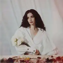 A woman with long dark hair wears loose-fitting white clothing and cradles a bunch of white roses in one arm.